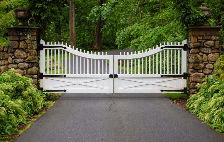 A white composite gate with custom black hinges sits between two dry-fit stone columns with metal lamps on top. There is a profusion of plants on either side of the driveway.