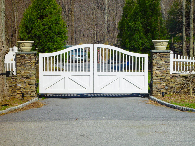 A white composite gate is half solid on the bottom and half made of vertical pickets on top. On either side there are stone columns with ornamental planters on top.