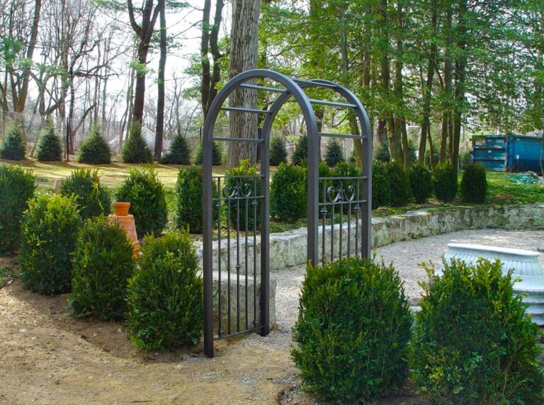 A wrought iron arbor frames a cut through a low stone wall between an enclosed gravel area on one side and bare dirt on the other. There are short evergreen bushes on either side of the arbor.