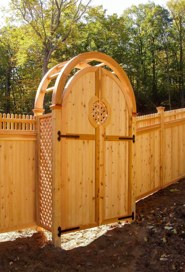 A wooden arbor is set into a solid wooden privacy fence. The fence is solid, with short pickets set on top. The arbor has latticed sides, an arched open top, and a solid gate.