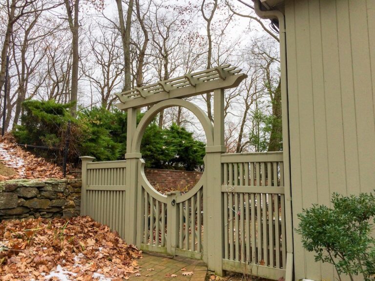 A gray-green painted picket fence spans a gap between a house of the same color and a short dry fit stone wall. A custom gate is set into the picket fence: the gate merges with an arbor whose arch is mirrored in a half-circle cutout at the top of the gate. A horizontal lattice is set on top of the arbor’s arch.