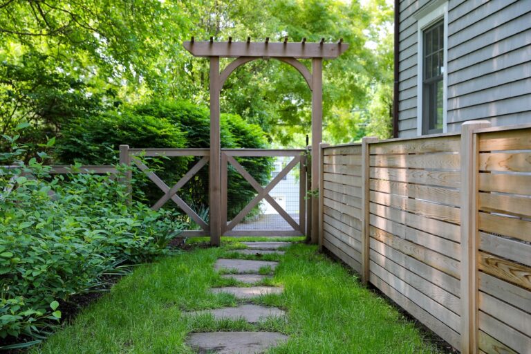 A simple square arbor and trellis frames a simple gate. On the right side of the gate is a low wooden fence with solid horizontal pickets, and on the left side of the gate is a hedge. Stepping stones lead towards the gate.
