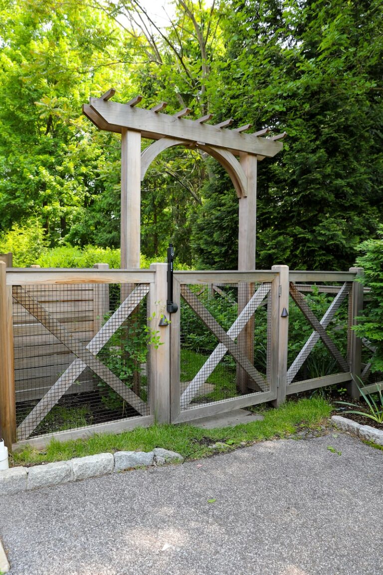 A simple wooden arbor sits behind a fence made of wood with x-shaped reinforcements and mesh. Both are in front of prolific green foliage.