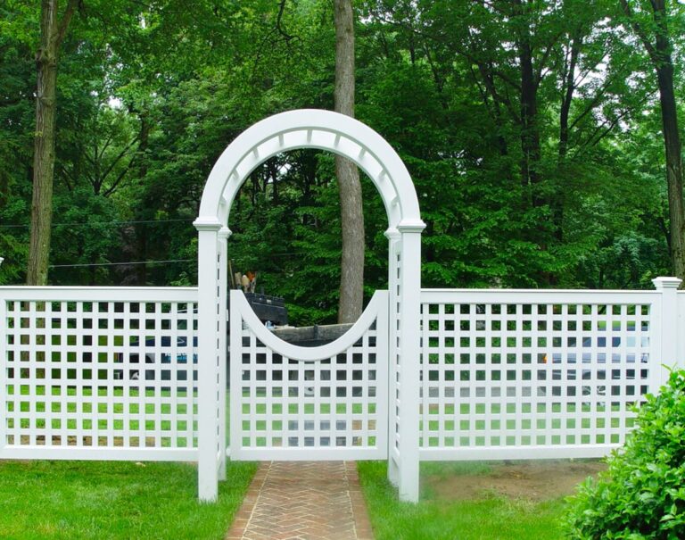 A white lattice fence has a white lattice arbor and gate. A brick path leads to the gate; beyond are mature trees.