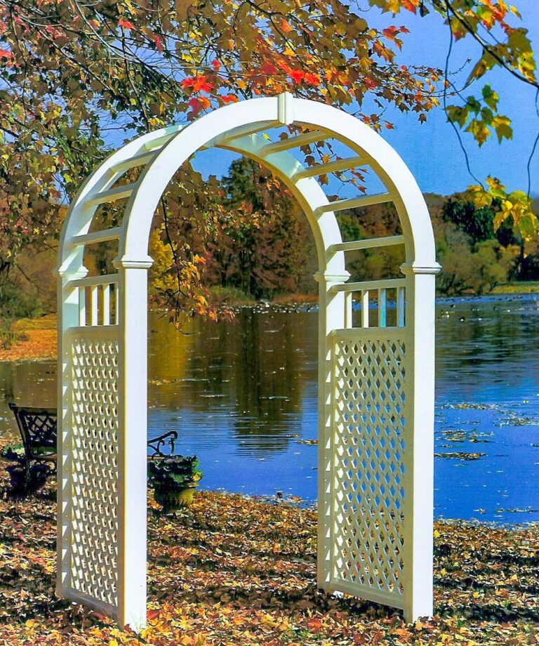 A freestanding white lattice arbor leads towards a wrought iron bench. The bench looks out over a lake. The trees around the lake are showing their fall colors, and there are leaves on the ground.