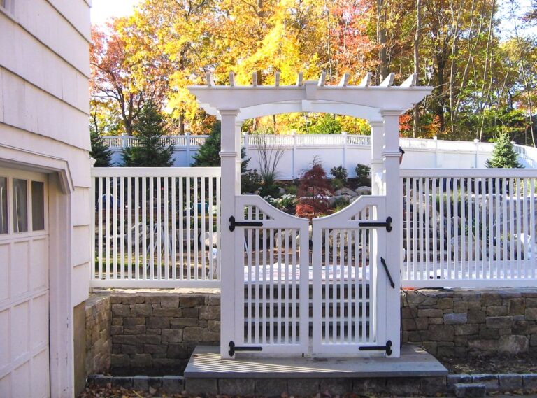 A stone retaining wall topped with a white picket fence is set back behind a white lattice arbor with a flat top. The arbor has a white gate with black metal hinges. Behind the fence is a carefully landscaped area with a mix of small trees and ornamental shrubs.