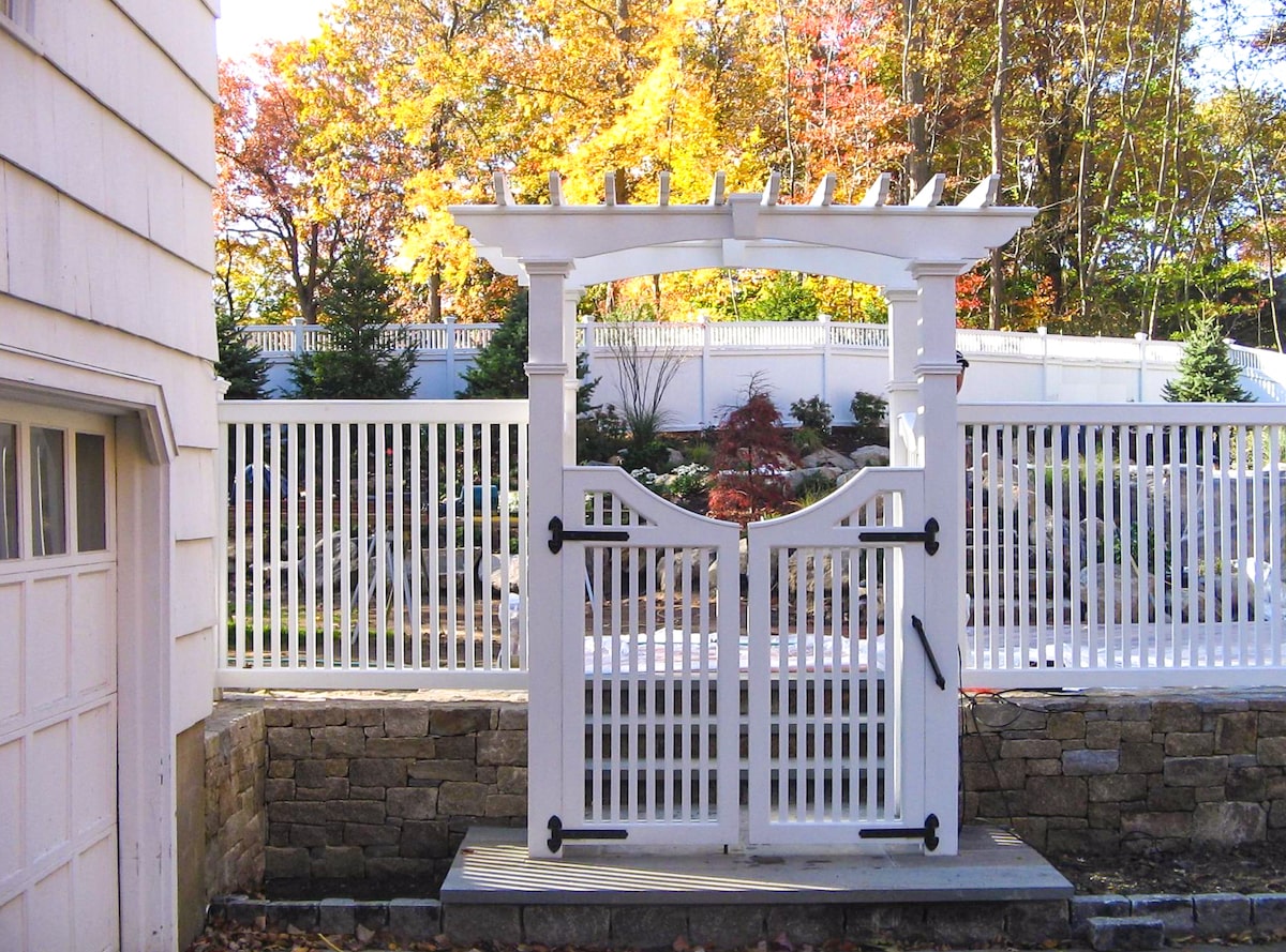 A stone retaining wall topped with a white picket fence is set back behind a white lattice arbor with a flat top. The arbor has a white gate with black metal hinges. Behind the fence is a carefully landscaped area with a mix of small trees and ornamental shrubs.