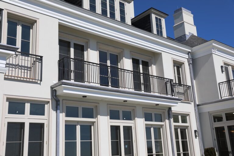 A medium-distance shot of a two-story white house with mullioned windows on both floors. A simple bronze balcony railing encloses the small balcony on the second floor.