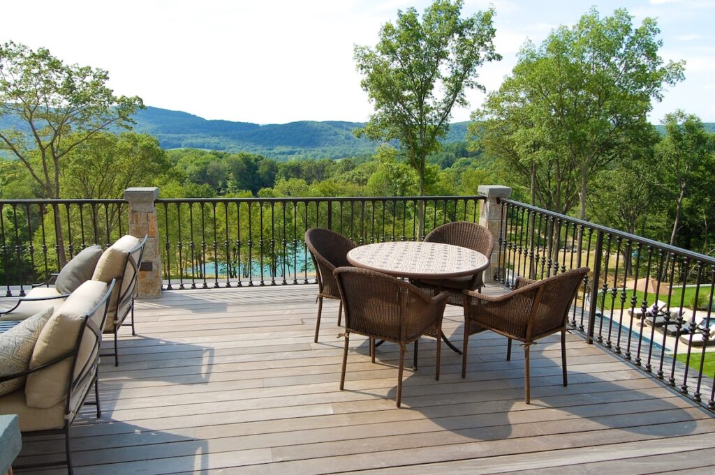 A deck overlooking the woods is adorned with a metal porch rail interspersed with stone pillars.