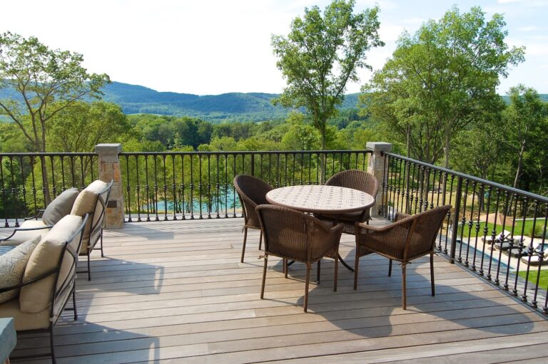 A short metal balcony railing with vertical spindles and stone pillars surrounds a protruding patio. There is deck furniture, including a round table and four chairs.