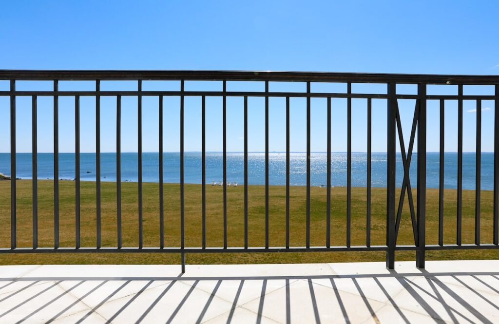 An ocean view is visible between the vertical spindles of a custom bronze railing.