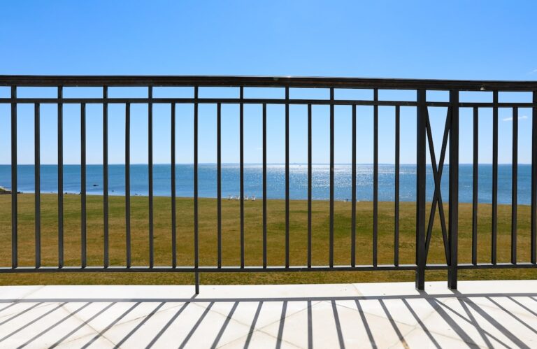 A minimalist metal balcony railing stands at the edge of a stone patio looking out toward the ocean.