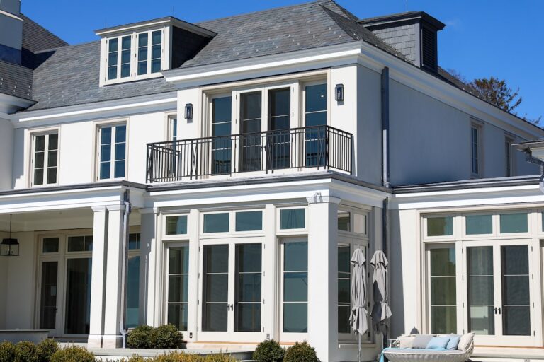 The viewer looks towards a white house with many windows, including glass French doors. Above the room with the French doors is a narrow balcony, surrounded by a simple black metal railing.