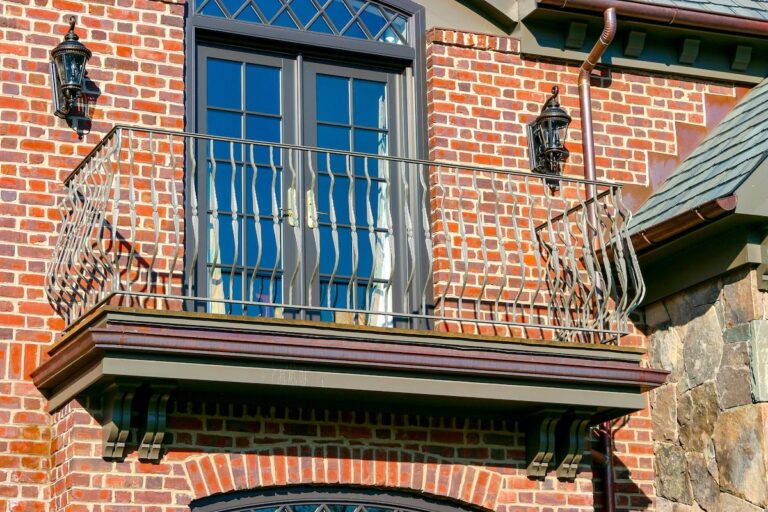 A set of glass French doors are set in a black frame on the second floor of a red brick house. They open out onto a small balcony protected by a gray metal balcony railing with curved spindles.