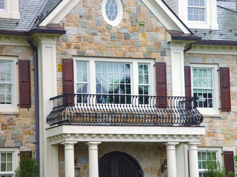 Each picket in this wrought iron balcony railing is dramatically curved, outward at the bottom and inward at the top, into an S-shape. The railing surrounds a faux balcony outside a large window on the second floor of a two-story stone house.