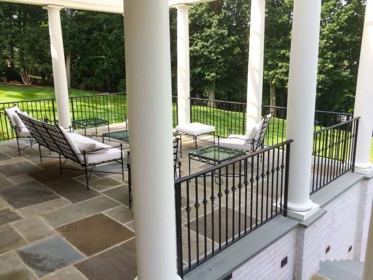 A multicolored flagstone patio protrudes over a green lawn. The patio is surrounded with simple wrought iron railings on three sides; the panels of the railing are framed by white columns that support the roof of the patio.