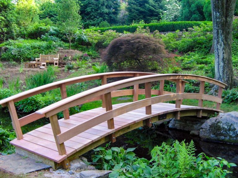 An arched garden bridge crosses the rocky banks of a stream in a lushly green garden. There’s a wooden bench in the background.