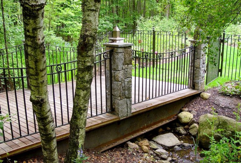 A flat garden bridge with a wooden base, stone columns, and wrought iron railings crosses a low creek bed.