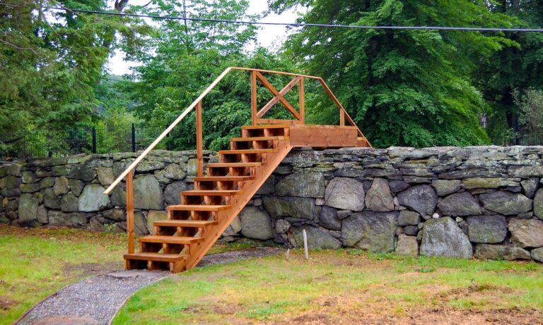 A wooden step bridge with a railing on one side leads up and over a stone wall.