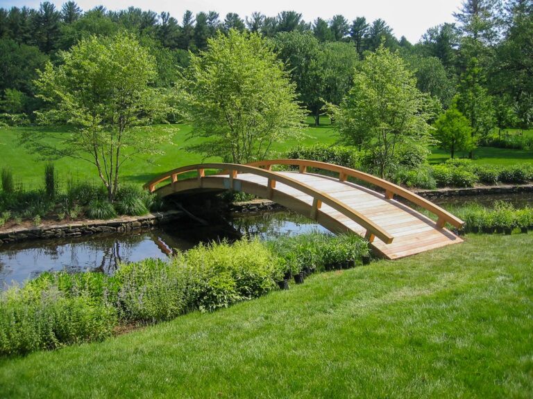 A wooden bridge stands out in a summer landscape. The pale brown and yellow color of the wood contrasts with the deep green of the surrounding trees and fields on either side of the stream it crosses.