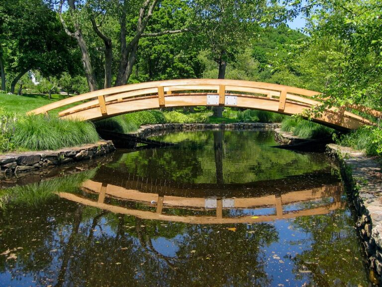 Viewed from downstream, this long, arched garden bridge passes over a wide stream with stone banks.