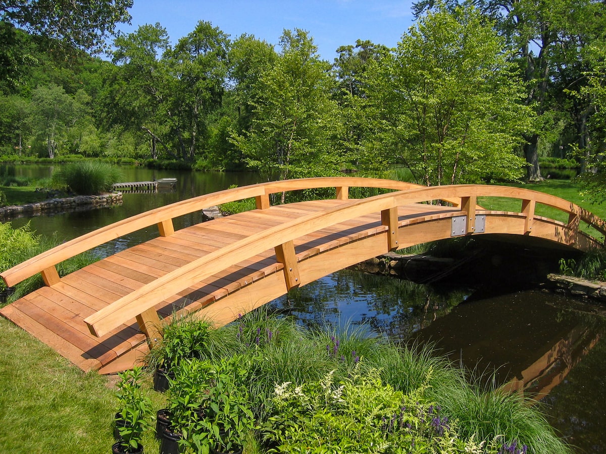 A long, wide, arched garden bridge spans the banks of a wide stream with reinforced stone banks and trees all around.