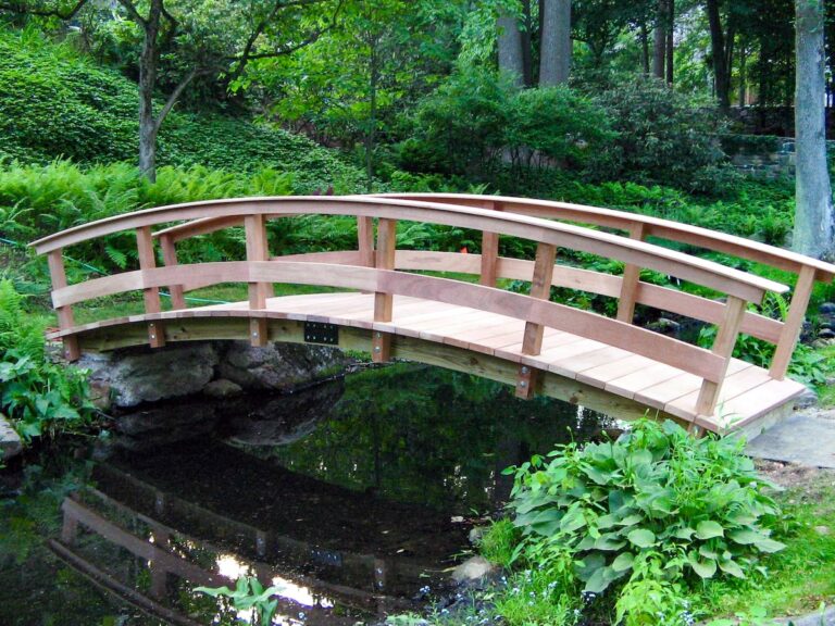 A long, arched garden bridge made from wood crosses from one bank to another, surrounded by greenery.
