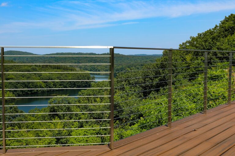 A tall cable railing is perched at the edge of a deck overlooking a wide expanse of woods with a river running through it.