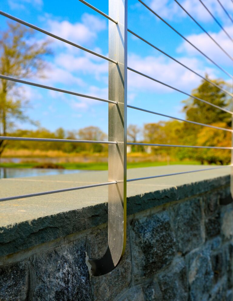A close-up image shows the curved base of a steel cable railing post that extends out from a stone wall.