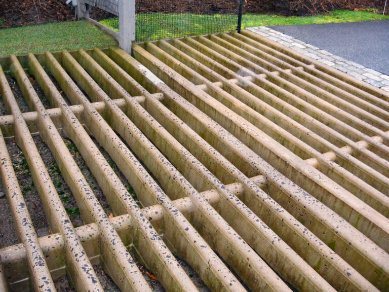 A close up shot of a concrete cattle guard set into an asphalt driveway. Bricks provide a transition from asphalt to concrete. The cattle guard is bordered on one side by grass, with mesh fencing separating the grass from the driveway.