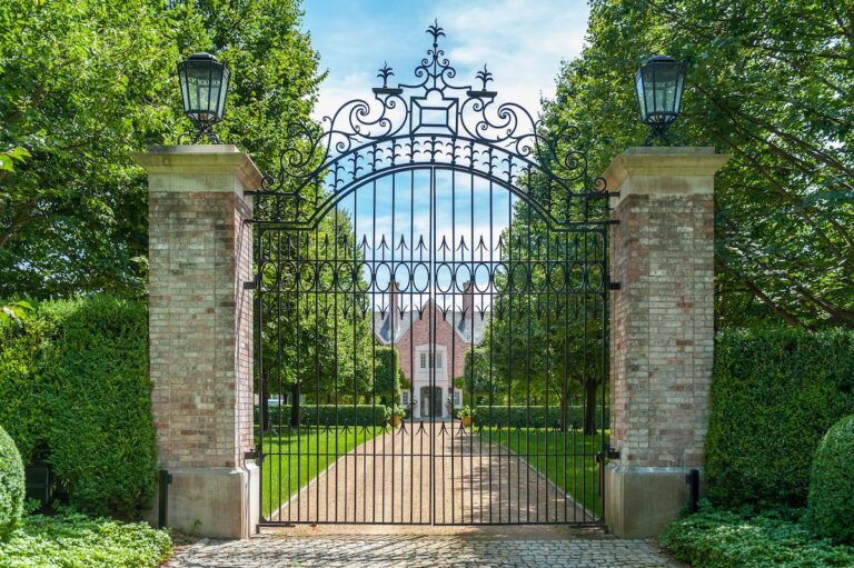 A tall wrought iron gate between two brick pillars protects a formal gravel driveway. There are wrought iron lamps on top of the pillars. The effect is imposing.