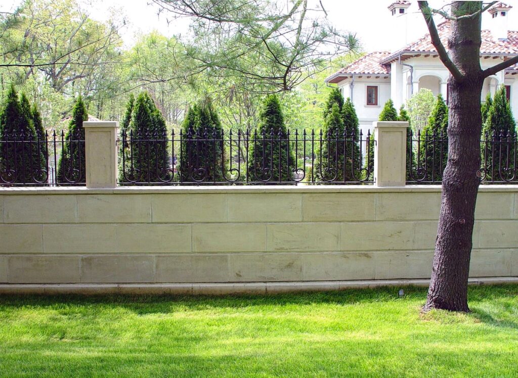 A solid light stone base is topped with widely-spaced wrought iron spindles. Evergreen shrubs behind the fence further screen a stucco house from view.