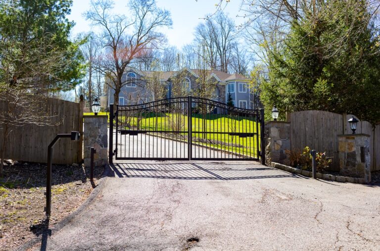 A simple metal double gate hangs between a wood panel fence with stone posts. A green lawn and large home are visible beyond the gate.