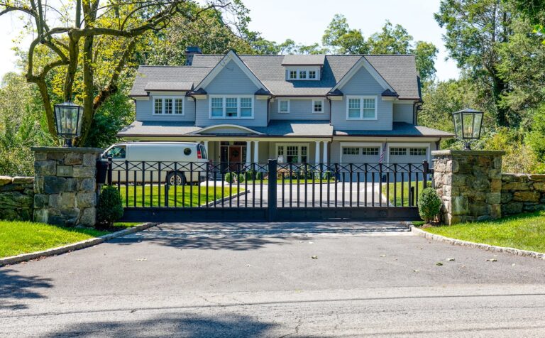 A simple short metal double gate sits between stone columns. A two-story blue home is visible beyond.