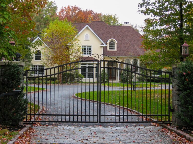 A metal swing gate with vertical spindles and S-shaped scrollwork guards a driveway leading to a 2-story yellow home.