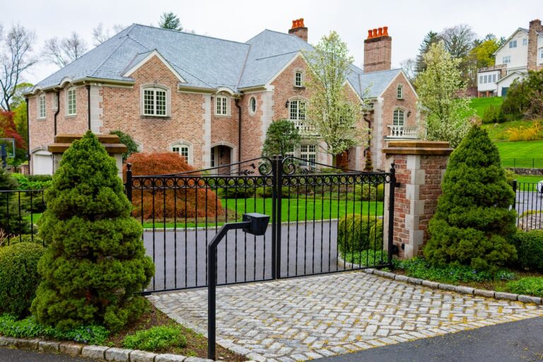 A metal swing gate between brick pillars with a call box in the foreground. A large and imposing brick house is visible beyond the gate.