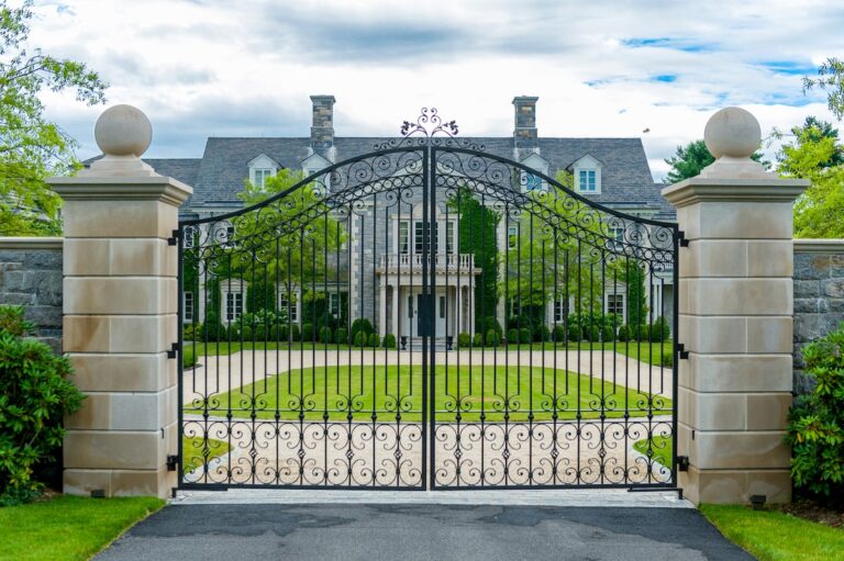 A wrought iron gate with curlicue ornaments framing the vertical spindles sits between two gray stone pillars. There are circular stone ornaments on top of the columns on either side.