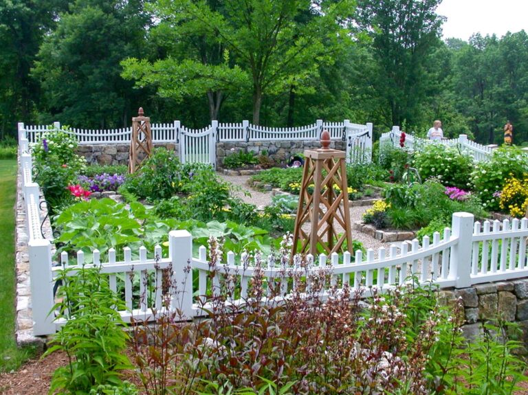 A fence made of a dry-fit stone base is topped with white picket panels. It surrounds a colorful garden laid out in square beds with gravel paths between them.