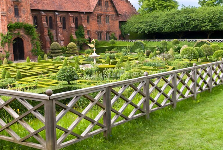 A formal English-style garden with shaped hedges is enclosed by a low wooden fence with diagonal pickets.