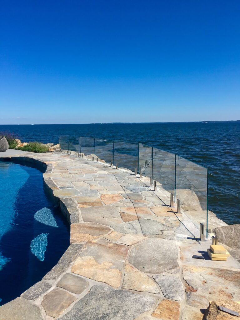 A glass railing atop a stone walkway divides a pool from the ocean.