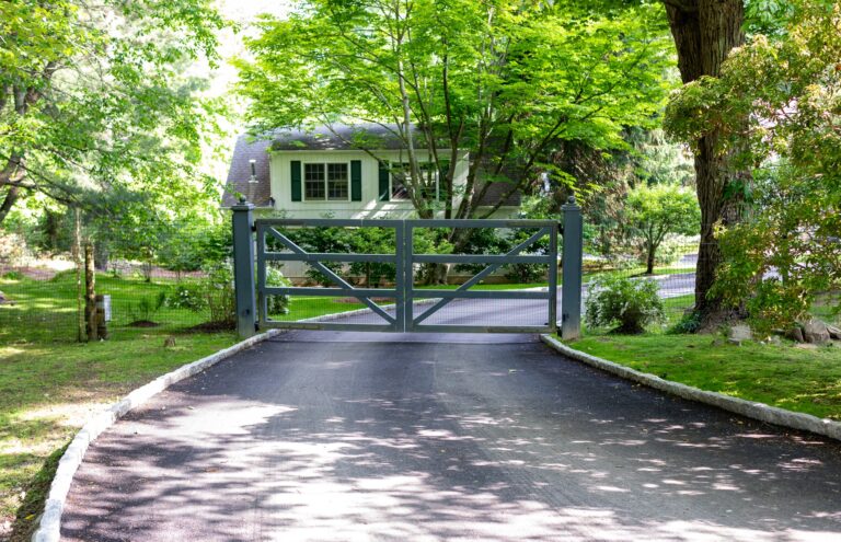 A post and rail wood gate is painted blue. It blocks a simple asphalt driveway leading between green lawn and mature trees.