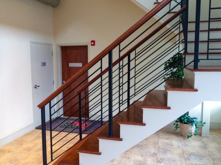 The interior railings on this wooden staircase feature dark-stained handrails that match the wood of the stairs with metal balusters and additional metal guardrails. Behind the railing is a wooden door leading to a law office.
