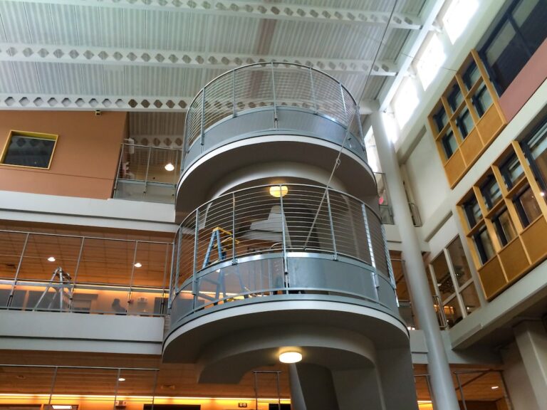Stainless steel interior railings enclose the circular stair landings on a multi-story interior stairway in the open lobby of a school.