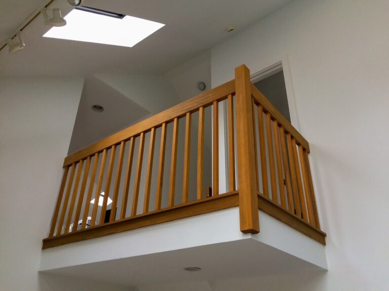 The camera looks up to a second-story interior landing protected by a half-height wooden interior railing. The vertical spindles are placed somewhat close together.