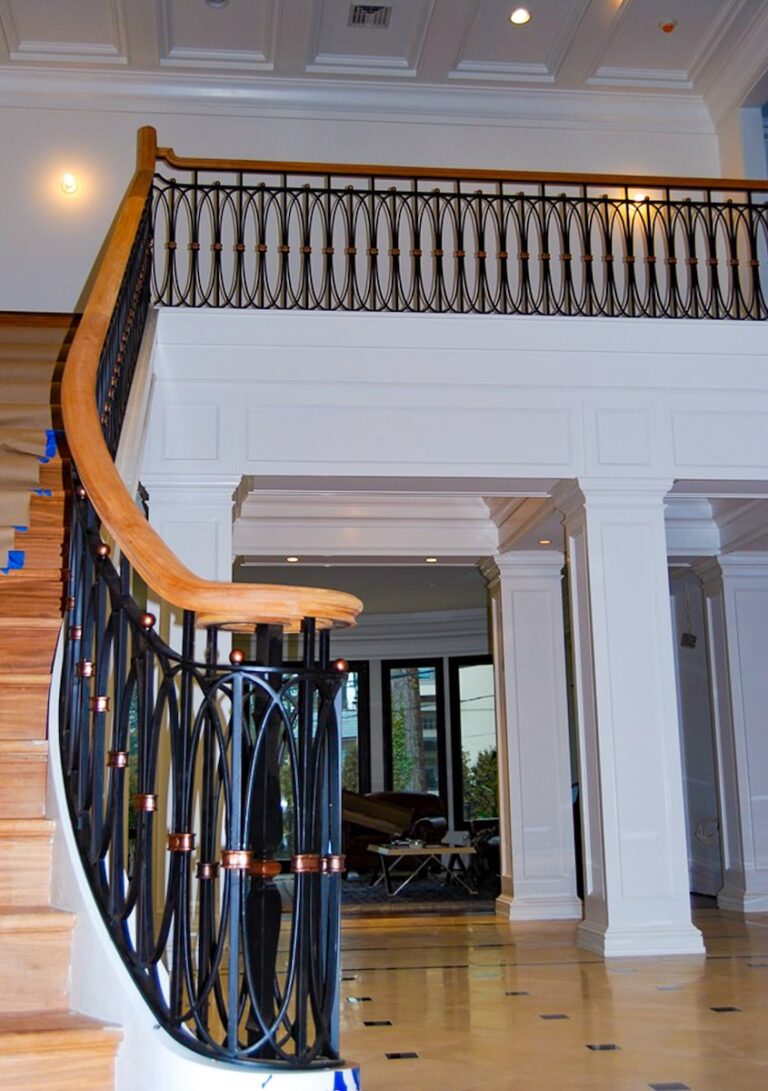 An ornate wood, wrought iron, and bronze interior railing runs along a balcony and down an imposing wooden staircase in a majestic home.