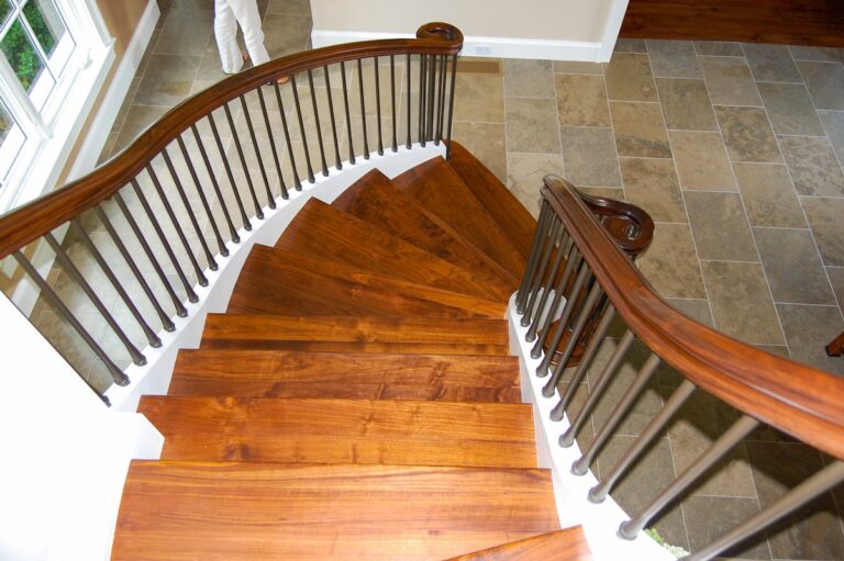 The viewer stands at the top of a wooden staircase looking down. The stairs curve slightly to the right, and the wooden handrail is supported by metal spindles.