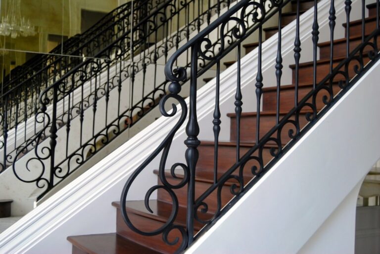 A close-up photo of ornate wrought iron S-shaped scrollwork at the bottom of an imposing dark wood staircase. The wrought iron railing is mirrored by the back wall.