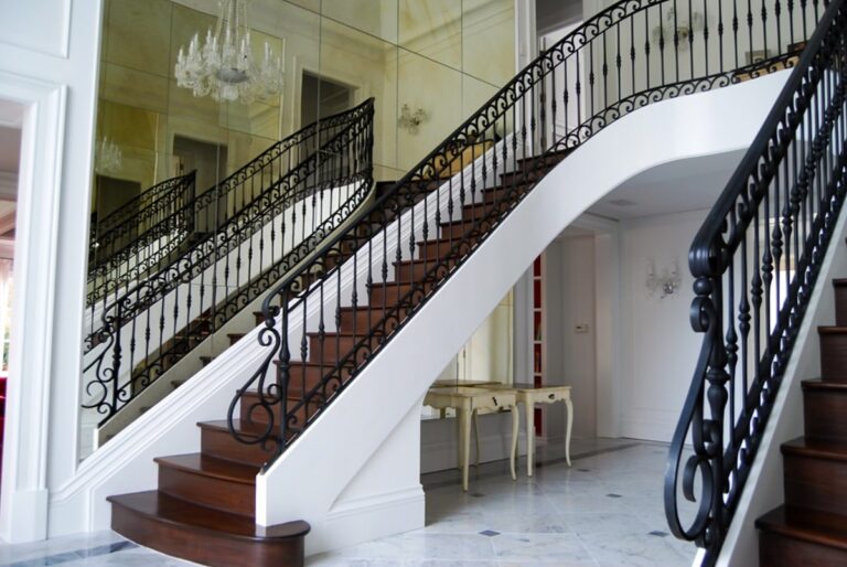 An ornate wrought iron interior railing frames an imposing dark wood double staircase in the entryway of a home. One wall beside the staircase is mirrored, doubling the staircase again.