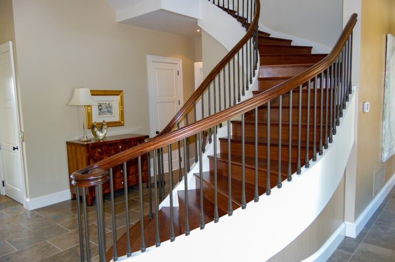 A staircase curves up and to the left in a home’s entryway. The stair treads are wood and match the handrail, while the closely-spaced spindles are metal.