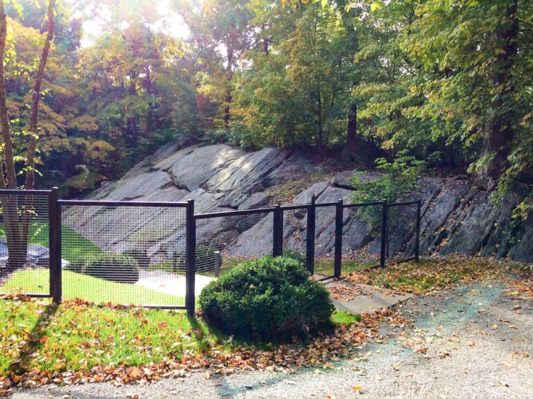 A black metal fence made of mesh panels divides a gravel area from a sunken wooded area.